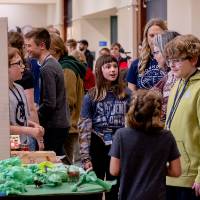Crowded hallway of a large building, where elementary school students talk with one another over trifold poster boards and dioramas, with parents and family around.